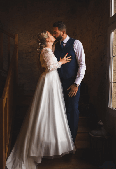 Photo d’un reportage mariage, photo où les mariés sont dans un escalier devant une baie vitrée leur donnant une lumière douce. Le couple s’embrasse. Elle est vêtue d’une longue robe fluide et lui d’un costume bleu avec veston. 