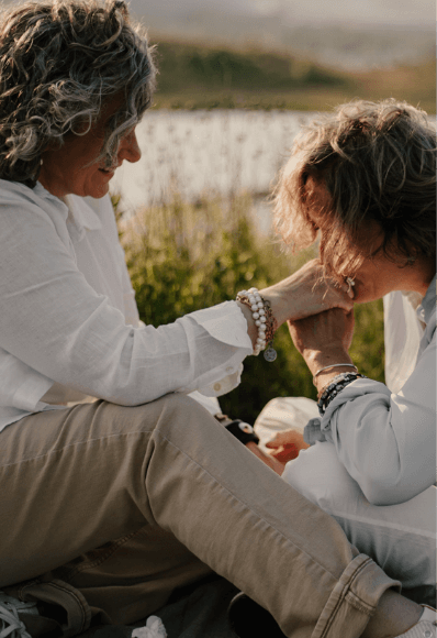 Séance de couple en extérieur de deux femmes d’âge mure. Un anniversaire de rencontre dans un lieu qui était cher à leur cœur. Assises sur la couverture de leur pique-nique, une des deux embrasse la main de l’autre. 