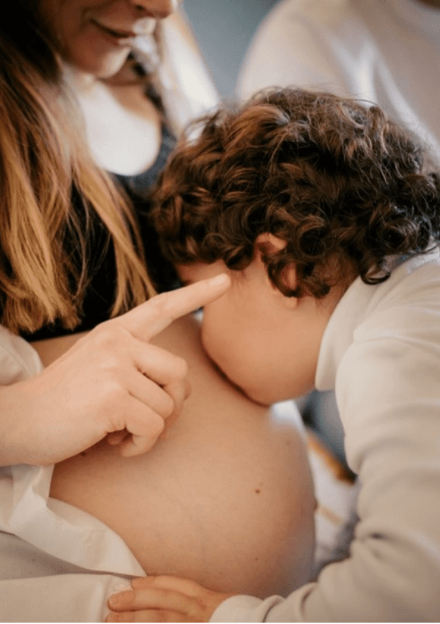 Séance photo de famille à la maison. Le fils de la maman âgé de 4 ans, embrasse le ventre rond de sa maman enceinte. 