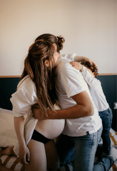 Séance photo de famille grossesse à la maison, dans la chambre des parents, les parents et l’enfant sont sur le lit se faisant un gros câlin. 