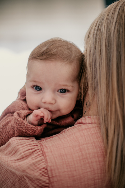 Photo prise par @sophiebacherephotos. Séance photo à la maison avec bébé. La photo montre le bébé porté par sa maman. Photo montrant le visage de bébé au-dessus de l'épaule de la maman.