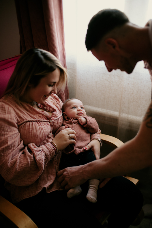 Photo prise par @sophiebacherephotos. Séance photo famille à domicile. La photo montre la maman assise tenant son bébé dans les bras tandis que papa est penché sur son bébé.