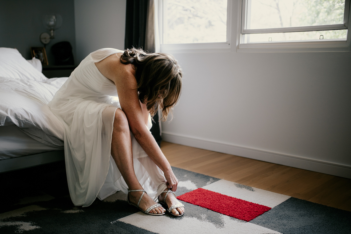 Photo prise par @sophiebacherephotos. Photo montrant la future mariée assise sur sont lit, laçant sa chaussure dans sa chambre sous la lumière tamisée de la fenêtre.