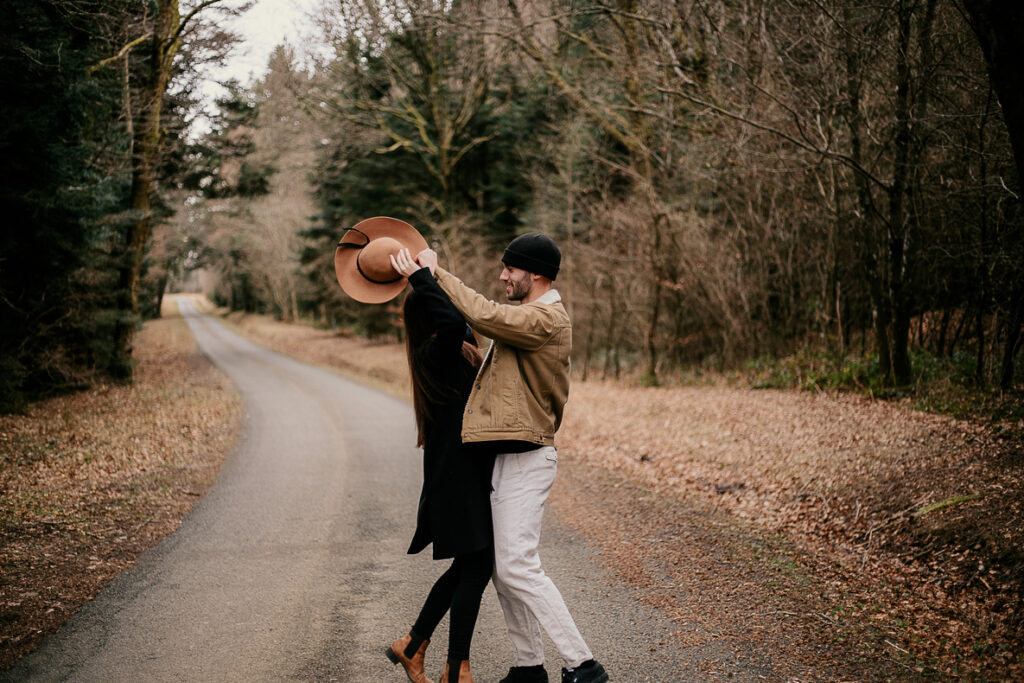 photo prise par @sophiebacherephotos. Séance photo de couple dans la forêt de la Montagne Noire dans l'Aude. La photo montre le couple sur une route entre la forêt. Ils s'amusent à s'attraper leurs couvre-chefs.