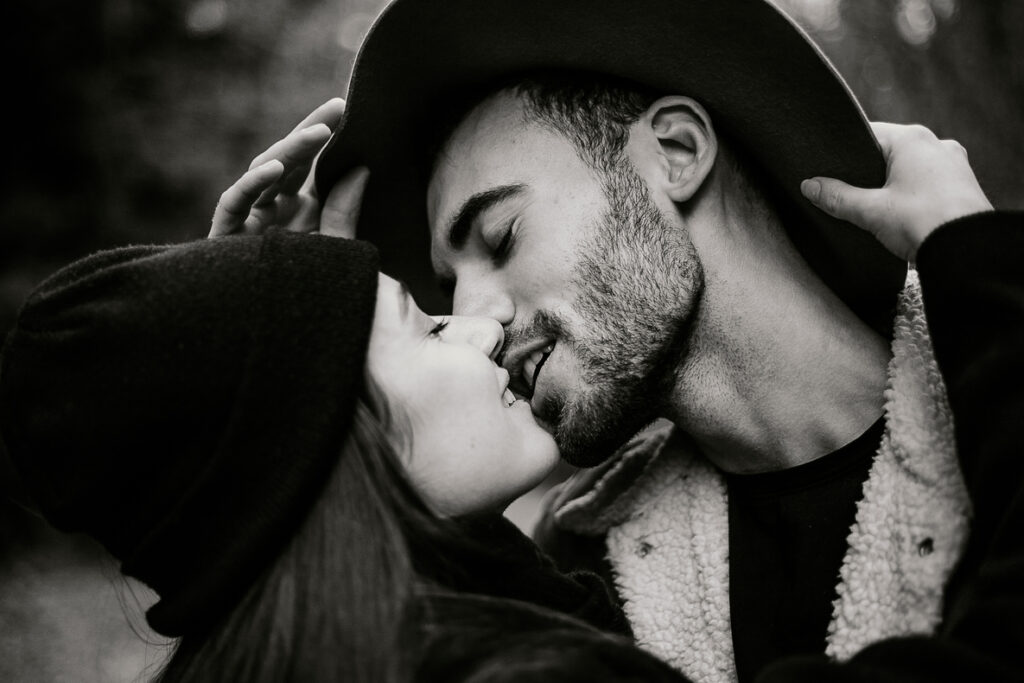 Photo prise par @sophiebacherephotos. Séance photo de couple dans la forêt de la Montagne Noire dans l'Aude. La photo en noir et plan montre un gros plan des visages du couple qui veulent s'embrasser, elle tient dans ses mains son chapeau qui est sur la tête de son homme.