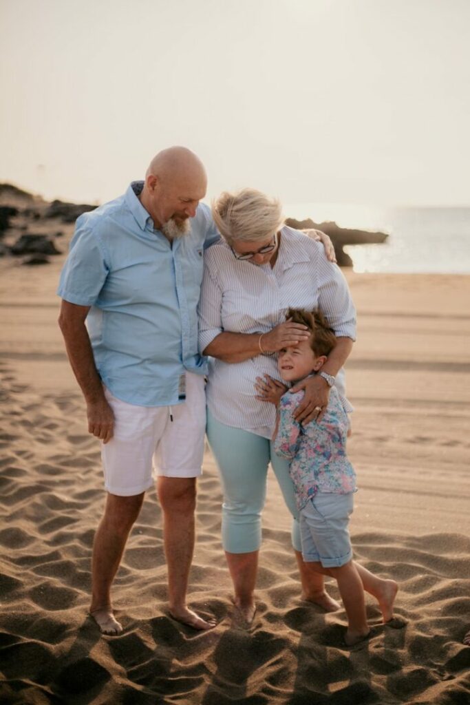 photo prise par @sophiebacherephotos. Séance photo de famille au bord de l'eau. La photo montre les grands-parents cote à cote sur la plage de Saint Pierre La Mer, la grand-mère dégage les cheveux du visage de son petit-fils collé contre ses jambes.