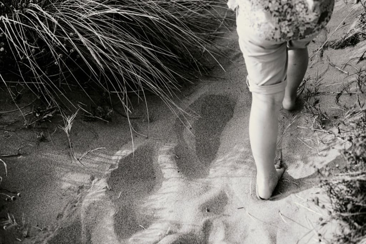 Photo prise par @sophiebacherephotos. La photo montre un enfant marchant dans le sable, on ne voit que ses jambes et la flore. Photo noir et blanc