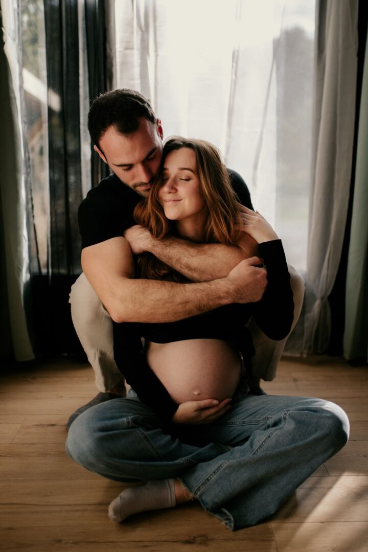 E&M-GROSSESSE-SOPHIE-BACHERE-PHOTOS-64 Photo prise par @sophiebacherephotos. Séance photo famille à domicile. La photo montre la maman assise tenant son bébé dans les bras tandis que papa est penché sur son bébé.